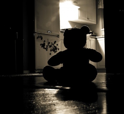 Small bear sitting in the dark, alone, on a kitchen floor.